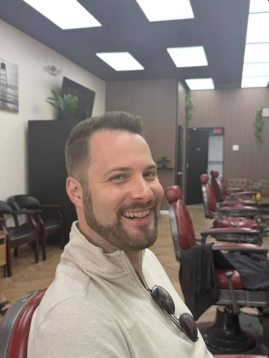 A smiling client with a short side part and a shaped beard, in one of the red barber chairs